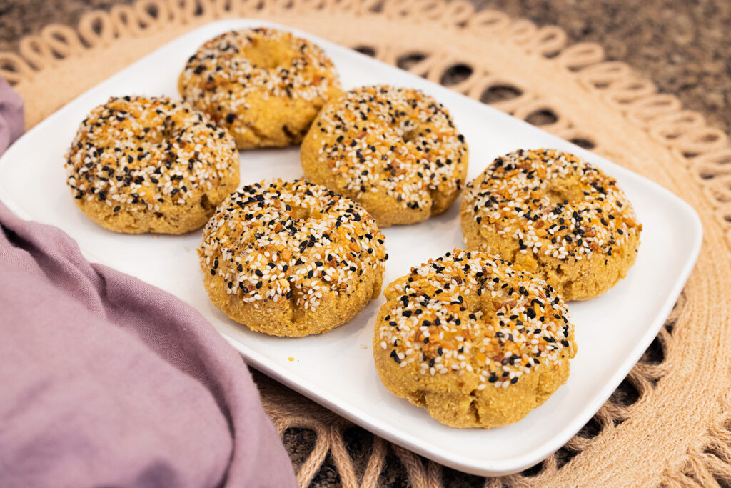 Close up landscape-style photo of a white platter filled with small everything bagels on a brown placemat with a purple napkin next to it.