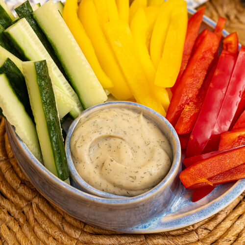 A closeup landscape-style photo of colorful cucumber and bell pepper slices in a blue ceramic dish with a large swirled dollop of the cannellini dip, all on a woven placemat.