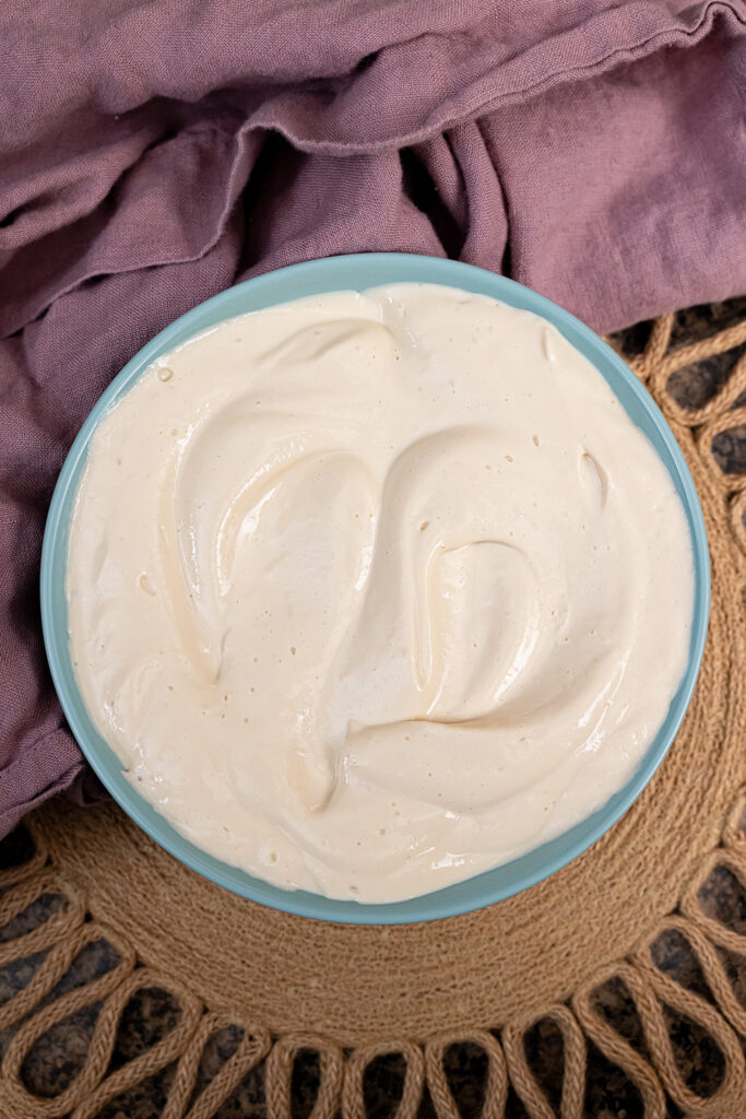 An overhead portrait-style photo of a blue bowl filled with tofu cream cheese on top of a brown placemat with a purple napkin next to it.