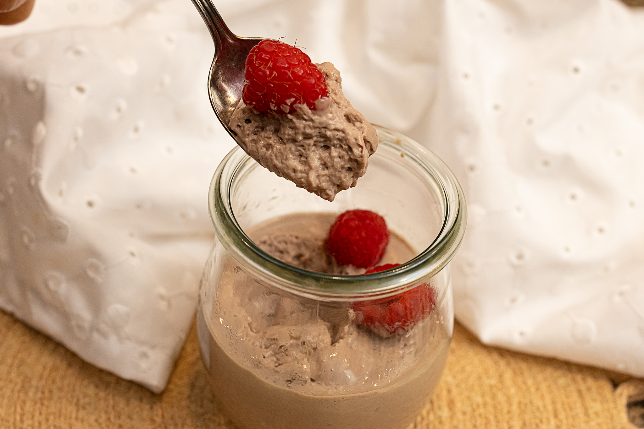 A landscape-style photo of a spoon hovering over a cup of chocolate tofu mousse on a tan woven placemat with a bit of the dessert and a raspberry in it, a white napkin in the background.