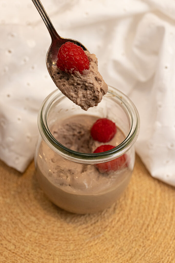 A portrait-style photo of a spoon hovering over a cup of chocolate tofu mousse on a tan woven placemat with a bit of the dessert and a raspberry in it, a white napkin in the background.