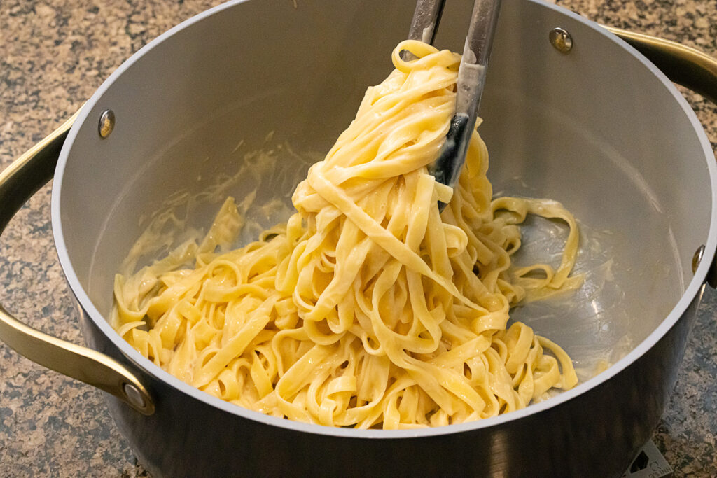 A landscape style photo of tongs twirling creamy fettuccine pasta in a gray pot on a brown granite countertop.