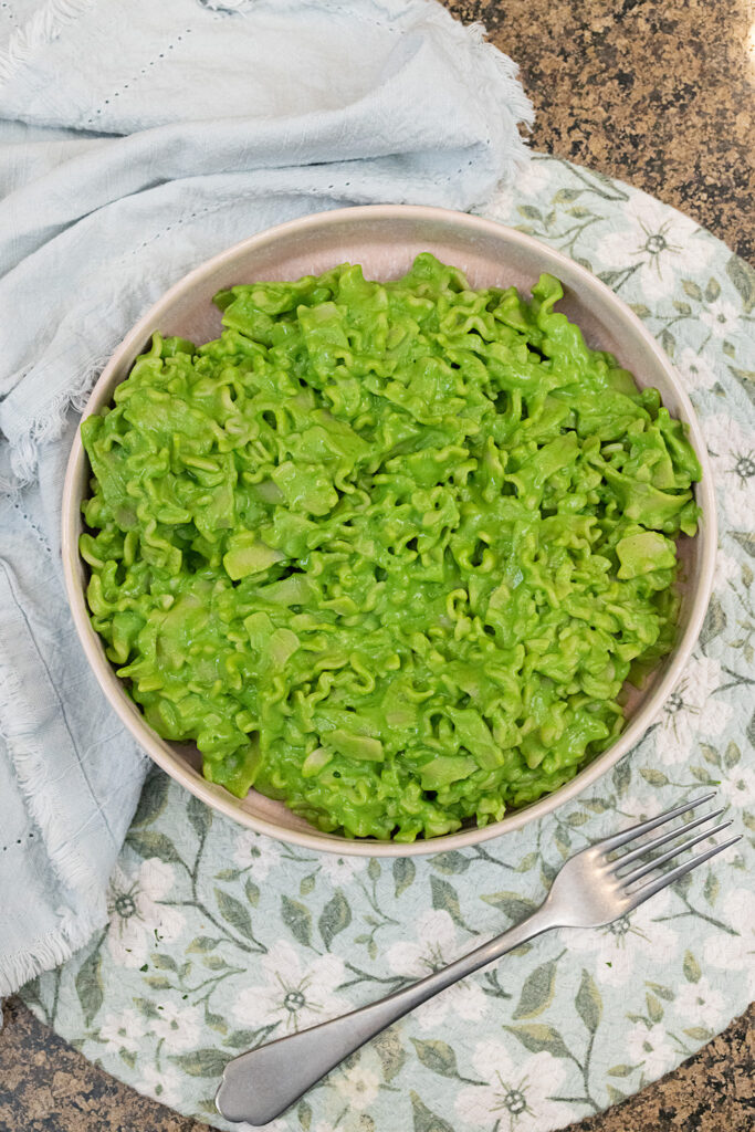An overhead portrait-style photo of a pink bowl of creamy green pasta on a blue floral placemat with a pewter fork.