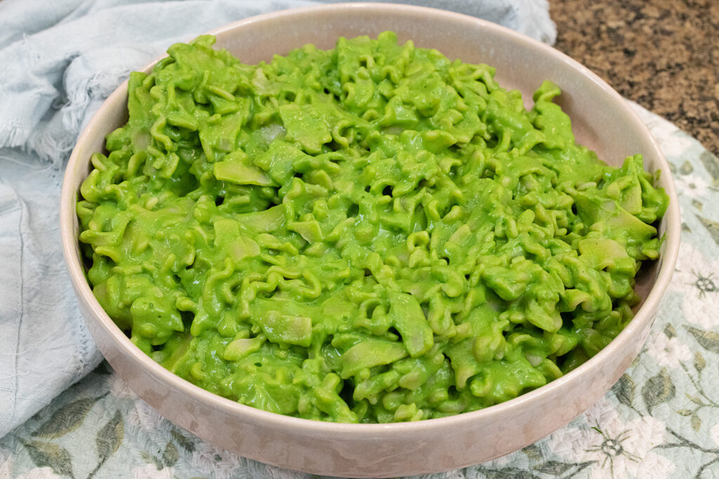 A 45 degree angle landscape-style photo of a pink bowl of creamy green pasta on a blue floral placemat.