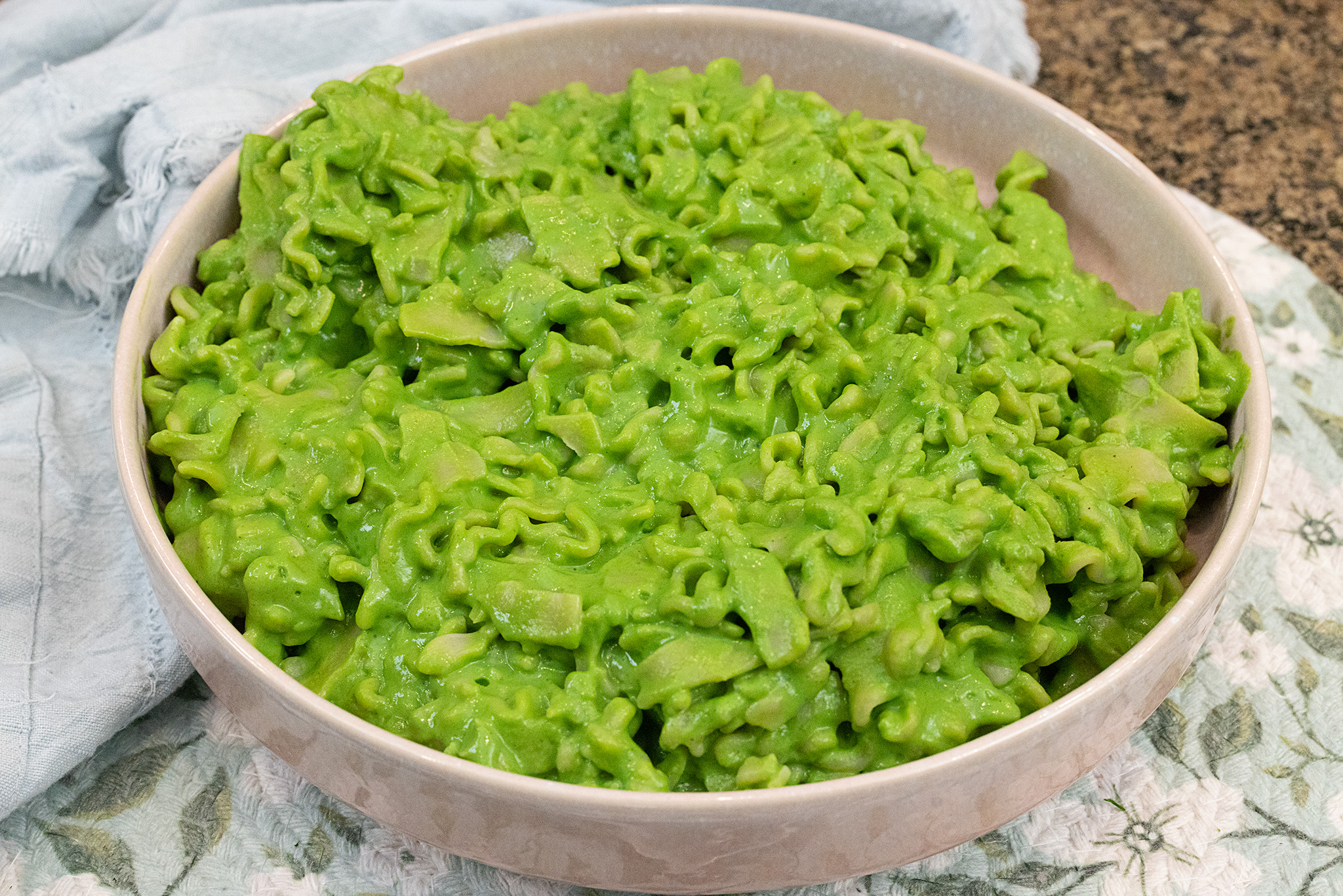 A 45 degree angle landscape-style photo of a pink bowl of creamy green pasta on a blue floral placemat.